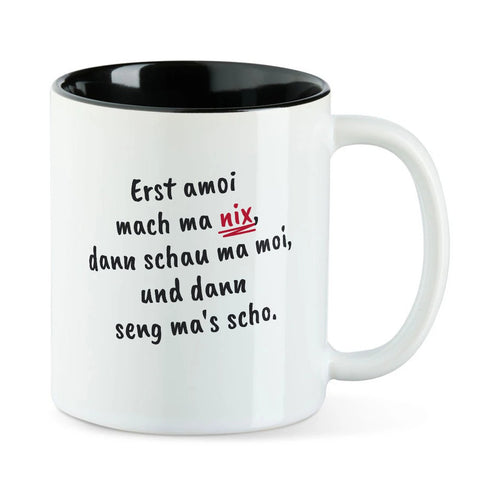 Weiße Tasse mit schwarzer Innenseite und dem bairischen Spruch „Erst amoi mach ma nix, dann schau ma moi, und dann seng ma's scho.“ in Pinselschrift.