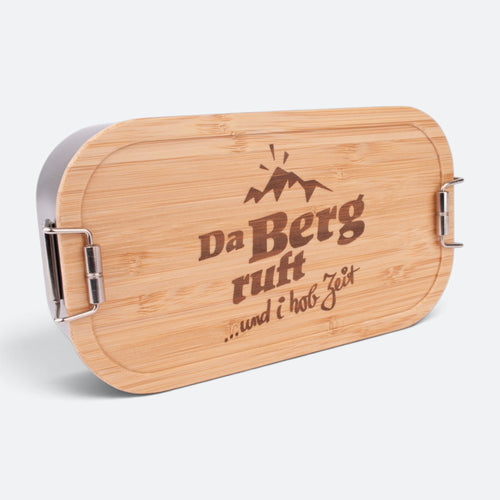 Brotzeitbox mit Bambusdeckel und der Gravur „Da Berg ruft … und i hob Zeit“ unter einem Berggipfel.