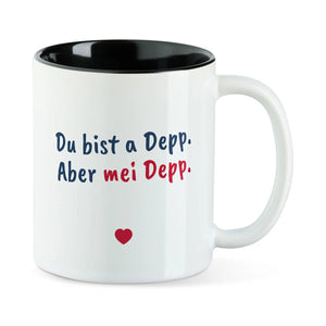 Weiße Tasse mit schwarzer Innenseite und dem bairischen Spruch „Du bist a Depp. Aber mei Depp.“ in Pinselschrift.