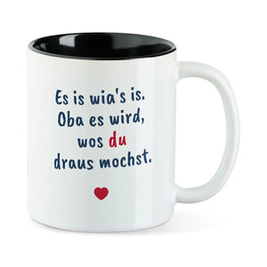 Weiße Tasse mit schwarzer Innenseite und dem bairischen Spruch „Es is wia's is. Oba es wird, wos du draus mochst.“ in Pinselschrift.