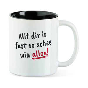 Weiße Tasse mit schwarzer Innenseite und dem bairischen Spruch „Mit dir is fast so schee wia alloa!“ in Pinselschrift.