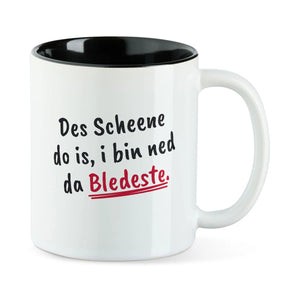 Weiße Tasse mit schwarzer Innenseite und dem bairischen Spruch „Des Scheene do is, i bin ned da Bledeste.“ in Pinselschrift.