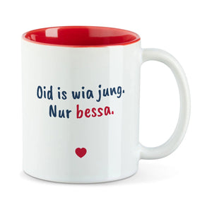 Weiße Tasse mit roter Innenseite und dem bairischen Spruch „Oid is wia jung. Nur bessa.“ in Pinselschrift.